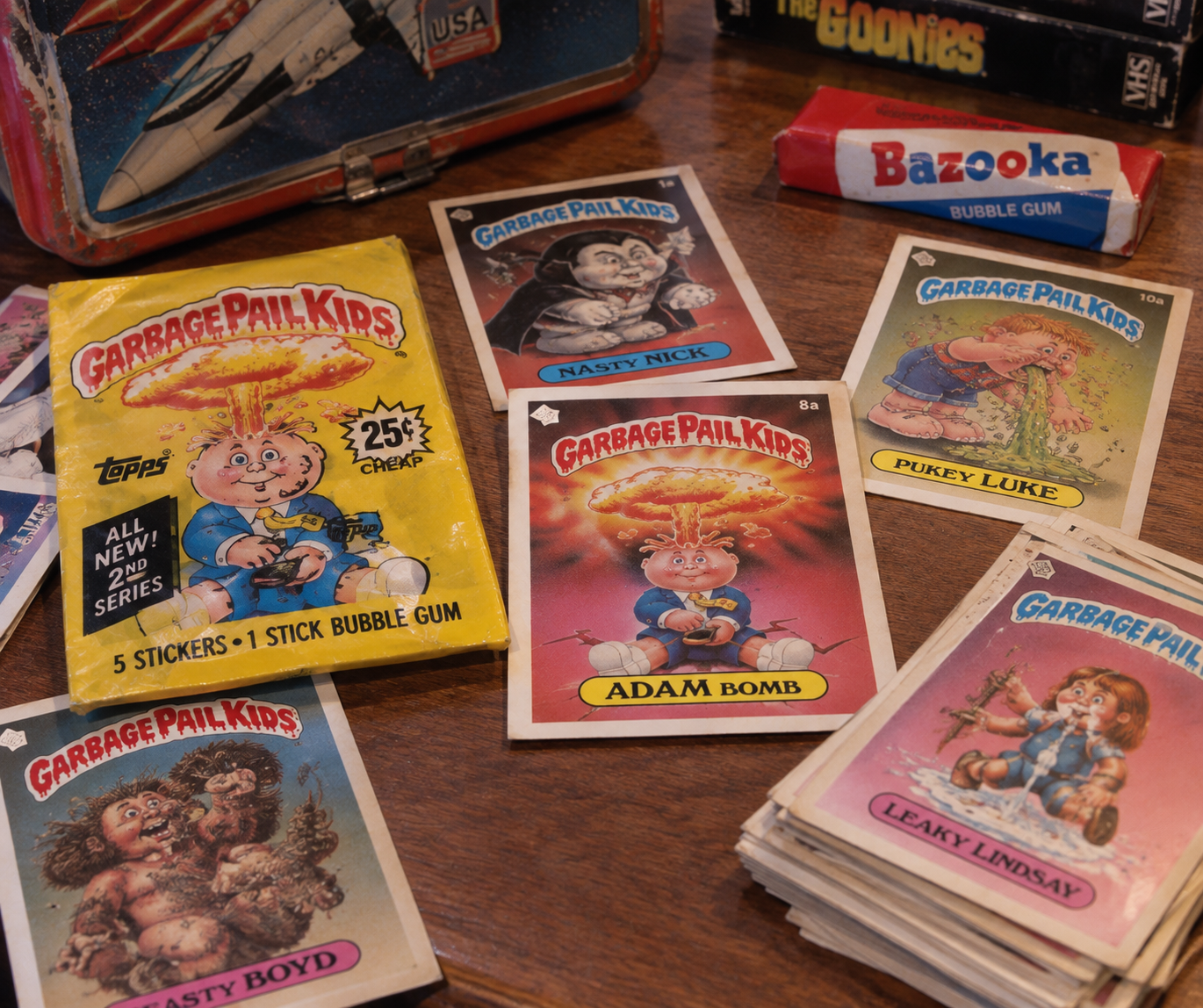 Several Garbage Pail Kids trading cards, including “Adam Bomb” and “Nasty Nick,” are spread on a table with a yellow pack, Bazooka gum, The Goonies VHS tapes, and a metal lunchbox in the background.