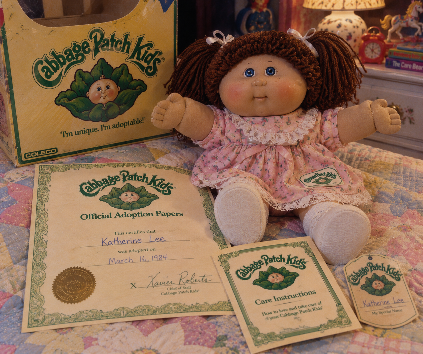 A Cabbage Patch Kids doll in a pink dress is displayed with its box, official adoption papers for Katherine Lee dated March 16, 1984, care instructions, and a birth certificate, all on a colorful quilt.
