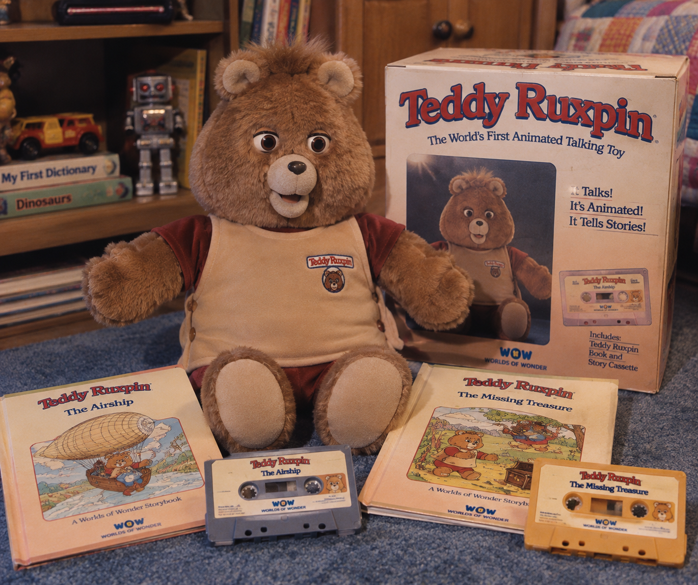 A Teddy Ruxpin talking bear plush sits with its box, storybooks, and cassette tapes on a carpeted floor. Vintage toys and books are visible in the background.