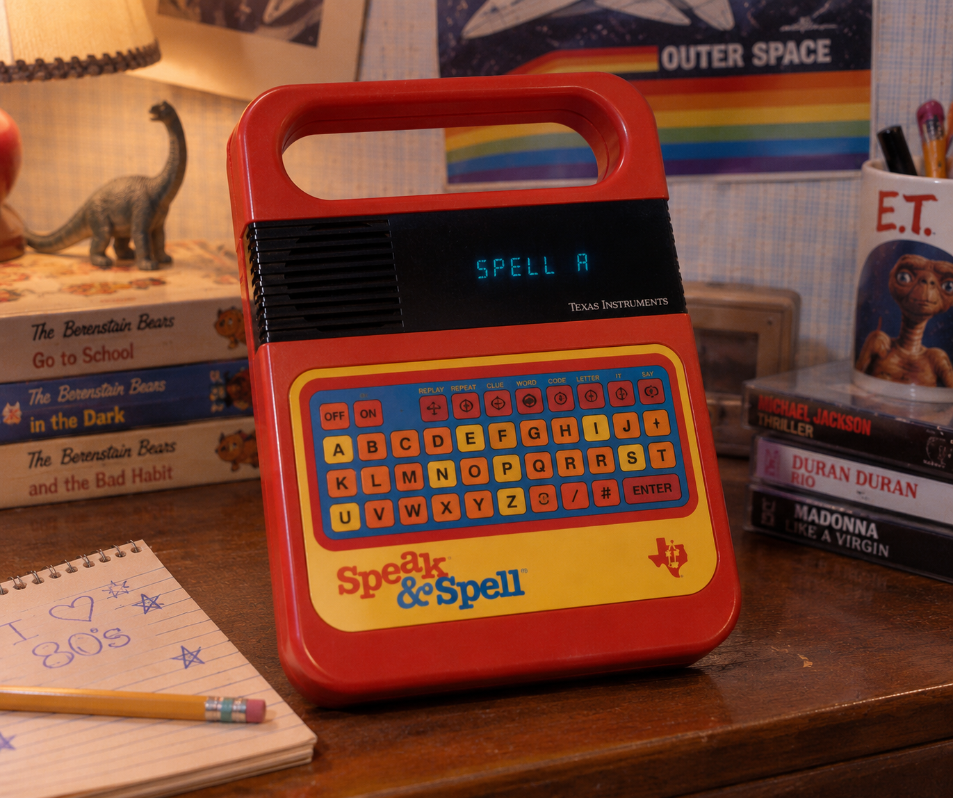 A Speak & Spell toy sits on a desk surrounded by books, a pencil, and a notebook. The screen displays “SPELL A.” A small dinosaur figure and 1980s memorabilia are in the background.
