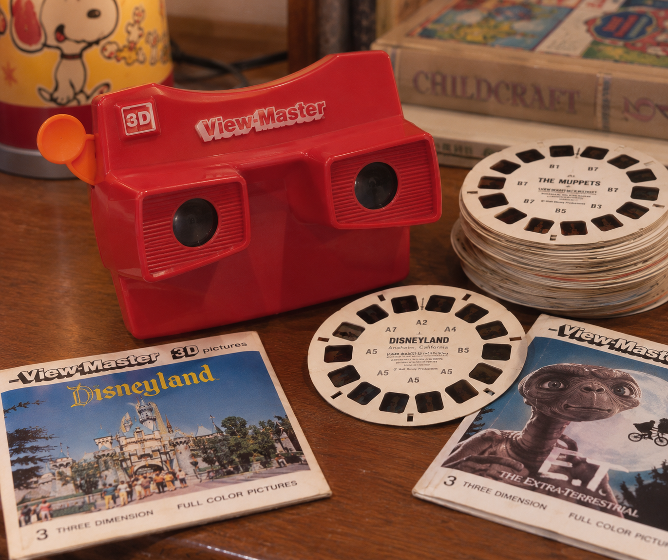 A red View-Master toy with reels and vintage packets for Disneyland, The Muppets, and E.T. sits on a wooden table next to books and a lamp. ’80s childhood essentials.