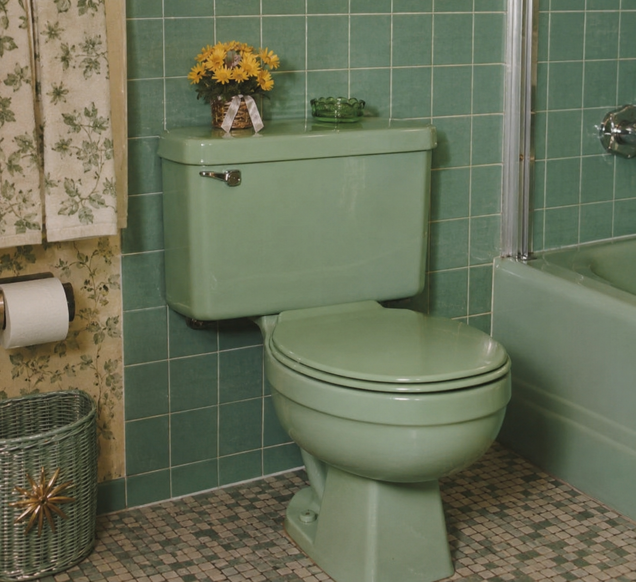 A vintage green toilet in a bathroom with green tiled walls, a matching bathtub, floral curtain, a wicker wastebasket, toilet paper, and a small flower arrangement on the toilet tank. 1960s bathroom.
