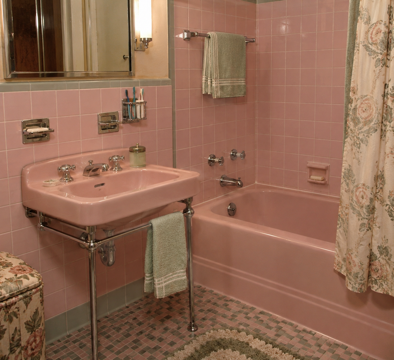 Vintage bathroom with pink sink, bathtub, and tiled walls. A towel hangs by the tub, floral shower curtain is pulled aside, and a green mat lies on the patterned tile floor. Mirror and toiletries are above the sink.