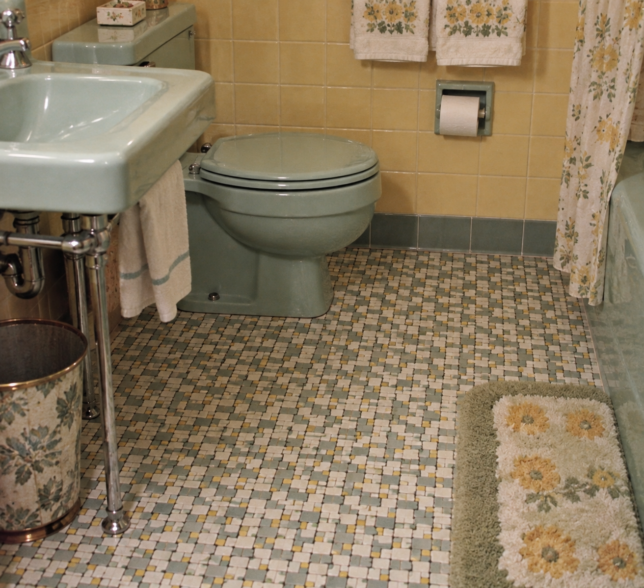 Vintage bathroom with green sink and toilet, yellow-tiled walls, mosaic tile floor, floral towels, a matching floral trash bin, and a coordinated bath mat featuring yellow and green floral patterns.