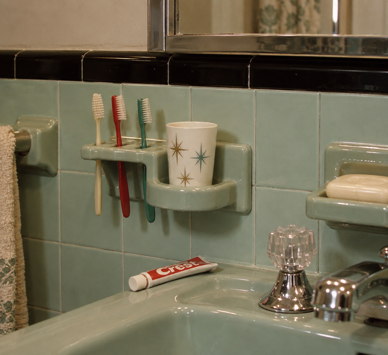 Three toothbrushes and a star-patterned cup sit in holders on a vintage mint green tiled bathroom wall, with a bar of soap, towel, faucet, and a tube of Crest toothpaste nearby.