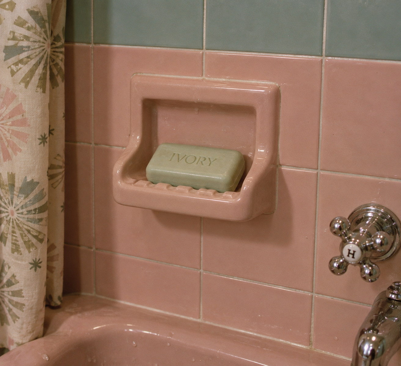 A bar of Ivory soap sits in a pink ceramic soap holder on a pink tiled bathroom wall, next to a vintage faucet marked “H” and a shower curtain with a retro pattern.