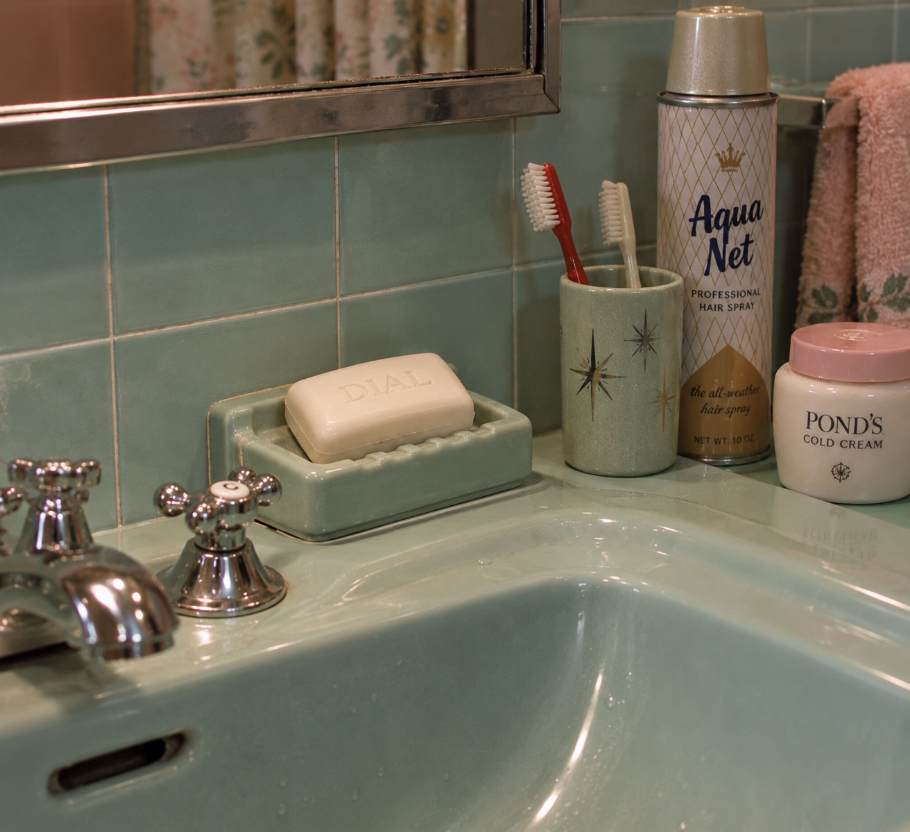 A vintage bathroom sink with green tiles, chrome faucet, a Dial soap bar, two toothbrushes in a starburst cup, Aqua Net hairspray, and Pond’s cold cream jar on the counter.
