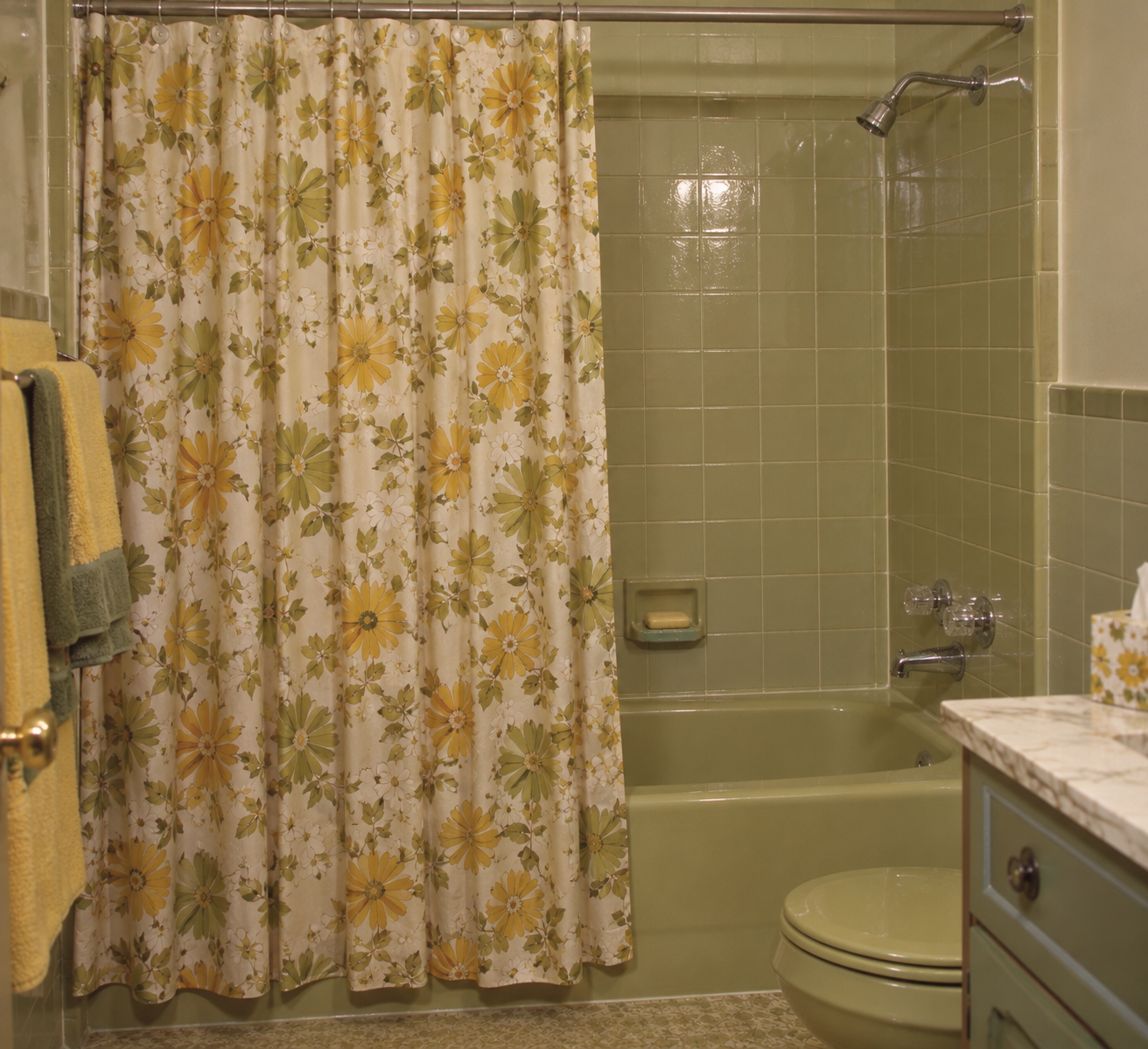 A vintage bathroom with green tiles, a green tub and toilet, a floral yellow and green shower curtain, yellow towels, and a marble countertop.