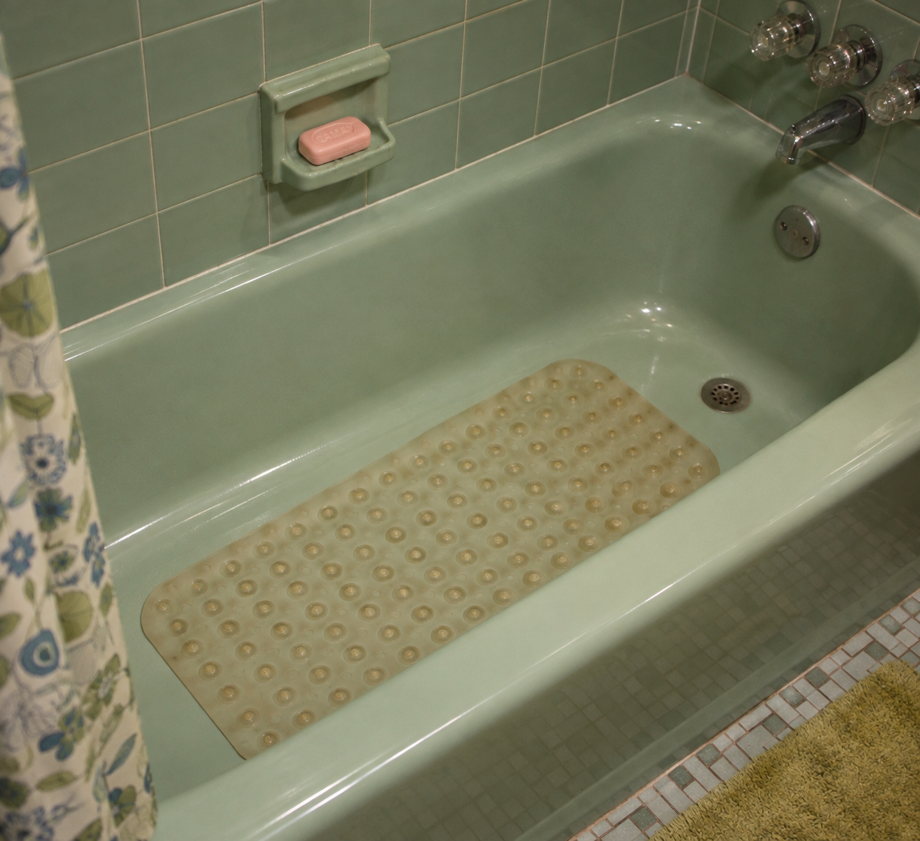 Green bathtub with a beige non-slip mat, green tile walls, silver fixtures, a bar of pink soap in a holder, a floral shower curtain, and a yellow bath mat on the floor.