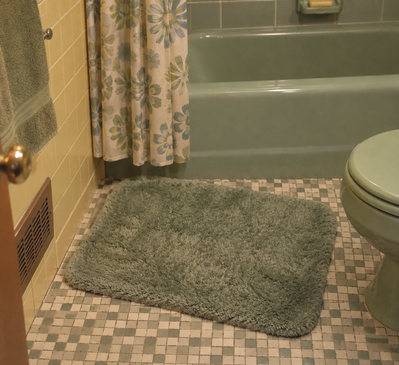 A green bath mat on a tiled bathroom floor next to a green toilet and bathtub, with a floral shower curtain and green towel visible.