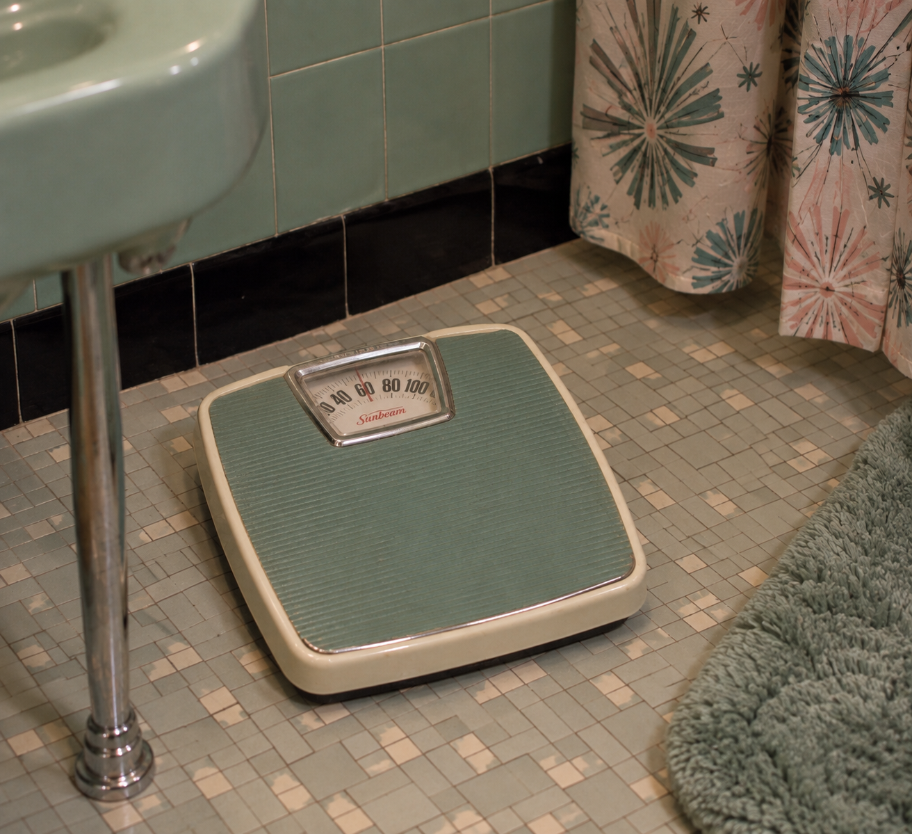 A vintage bathroom scale sits on a green-tiled floor beside a green bath mat, green sink, and retro-patterned shower curtain in a mid-century style bathroom.