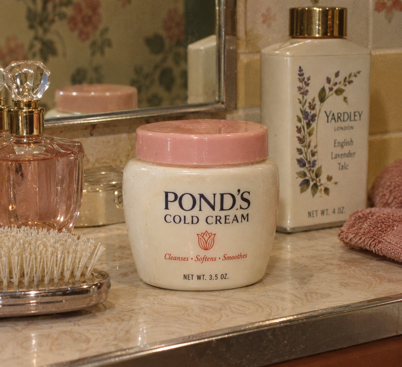 A jar of Pond’s Cold Cream sits on a bathroom counter near a perfume bottle, a hairbrush, a pink towel, and a bottle of Yardley English Lavender talc. A mirror and floral wallpaper are in the background.