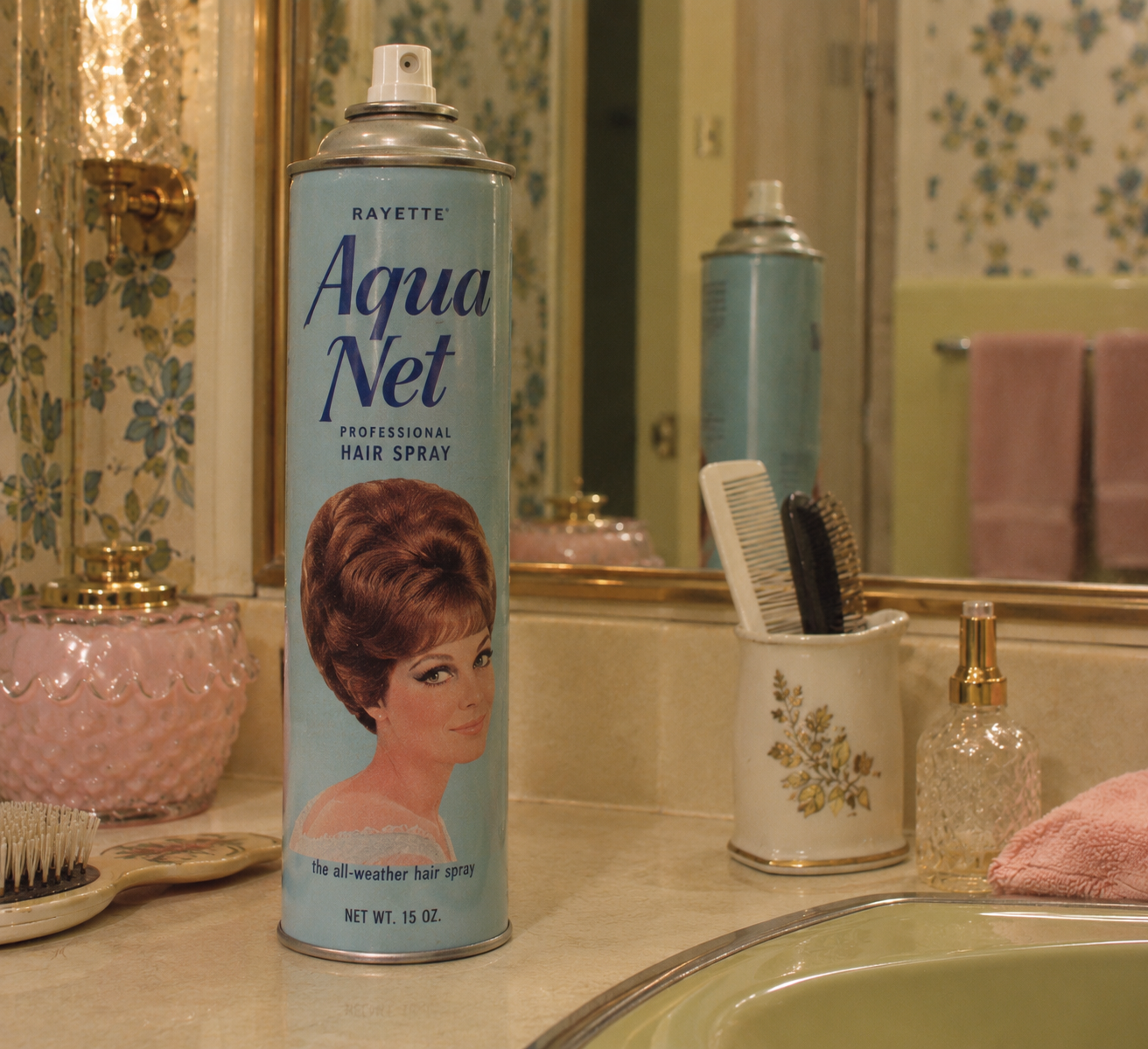 A can of Aqua Net hairspray sits on a bathroom counter near a sink, with a ceramic container holding combs and brushes. The bathroom has floral wallpaper, a mirror, and pink towels in the background.