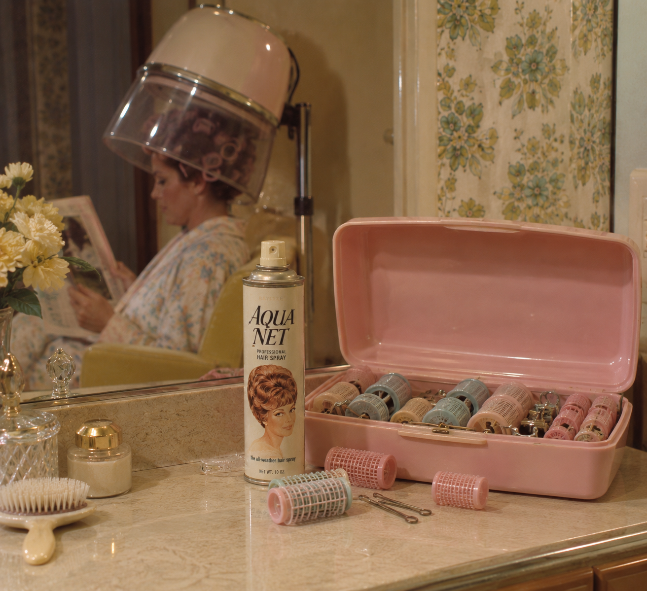 A woman sits under a hair dryer reading. On the counter are a can of Aqua Net, a pink box of hair rollers, a brush, pins, plus yellow flowers and toiletries. The room has floral wallpaper and a vanity mirror.