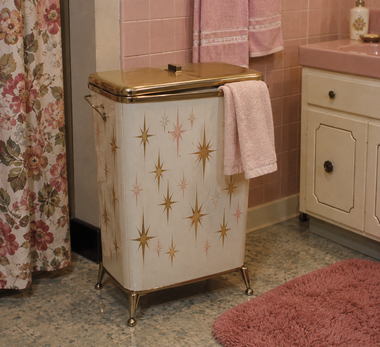 A vintage hamper with gold starburst patterns stands in a pink-tiled bathroom, topped with a pink towel. A floral shower curtain and pink rug add to the retro decor.