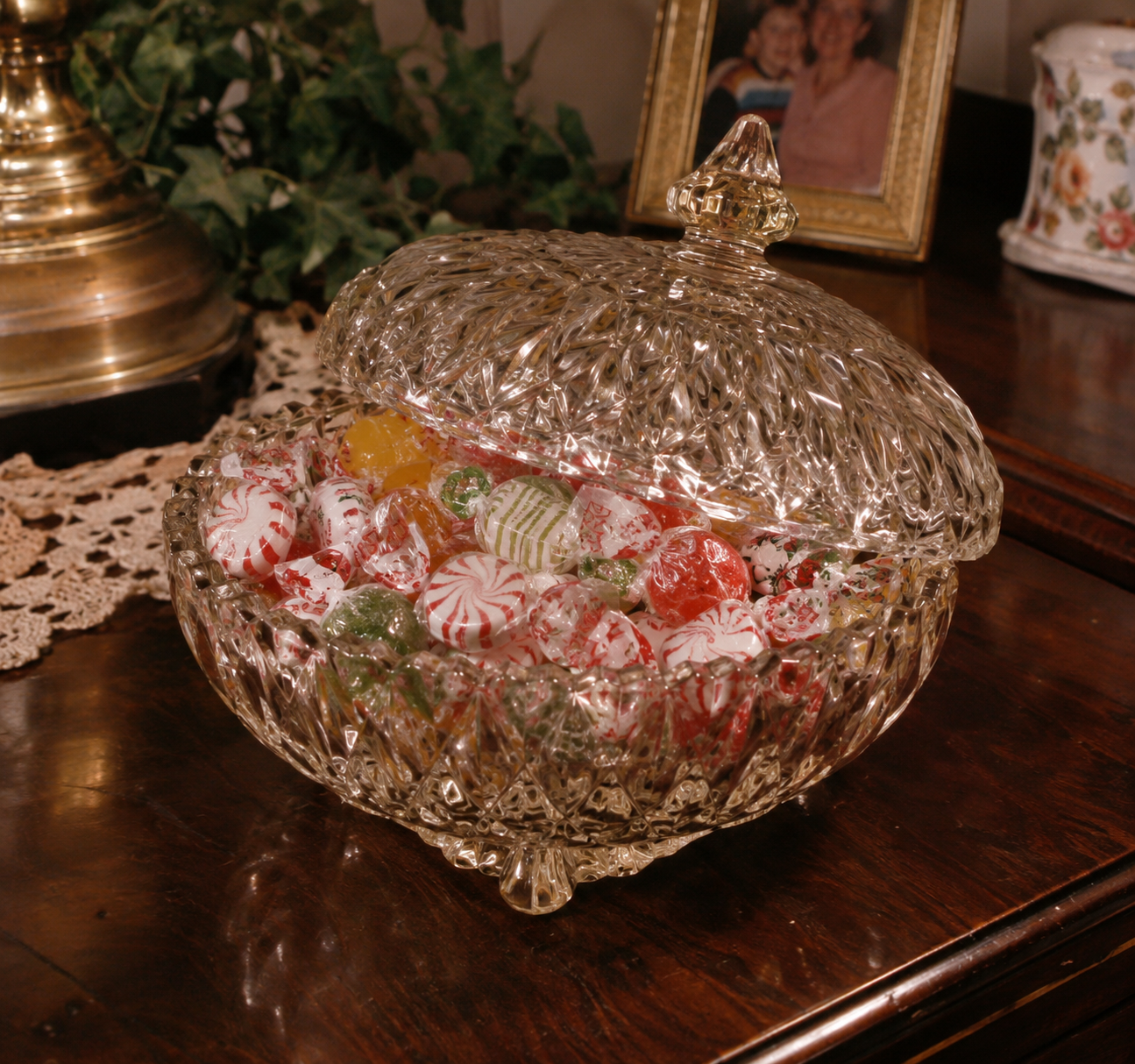 A crystal candy dish filled with assorted wrapped and unwrapped candies sits on a wooden table near a lamp, a framed photo, and a decorative plant.