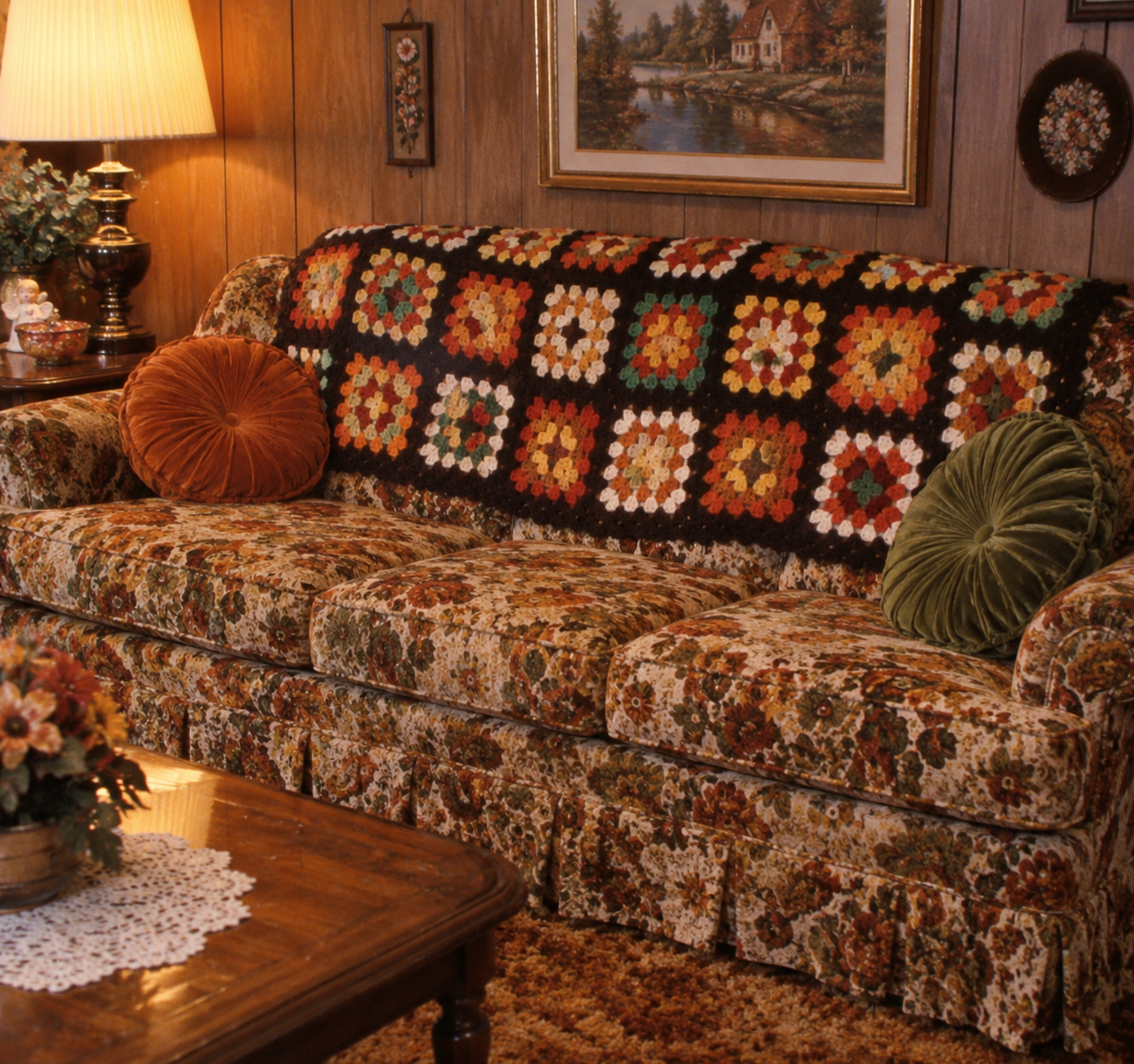 A vintage floral sofa with a colorful crocheted afghan and round pillows sits in a wood-paneled living room, with a lamp, framed art, and a flower arrangement on a wooden coffee table.