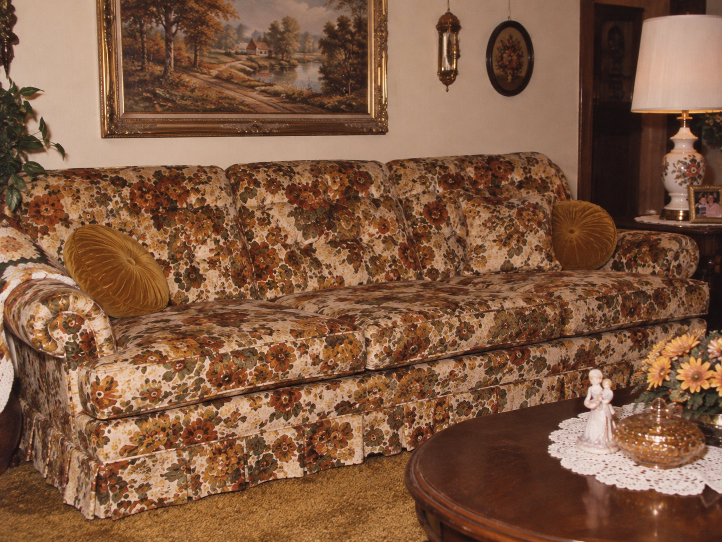 A vintage floral-patterned sofa with two round pillows sits in a cozy living room, decorated with framed art, a lamp, a doily, and a figurine on a wooden coffee table with flowers.