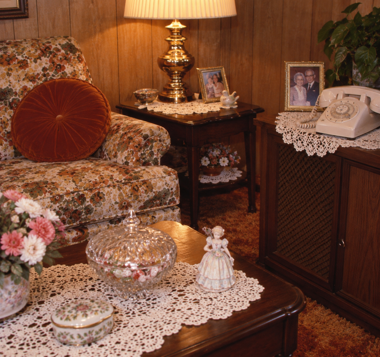 A vintage living room with floral furniture, lace doilies, a rotary phone, framed photos, a lamp, flowers, and decorative figurines on dark wood tables against wood-paneled walls.