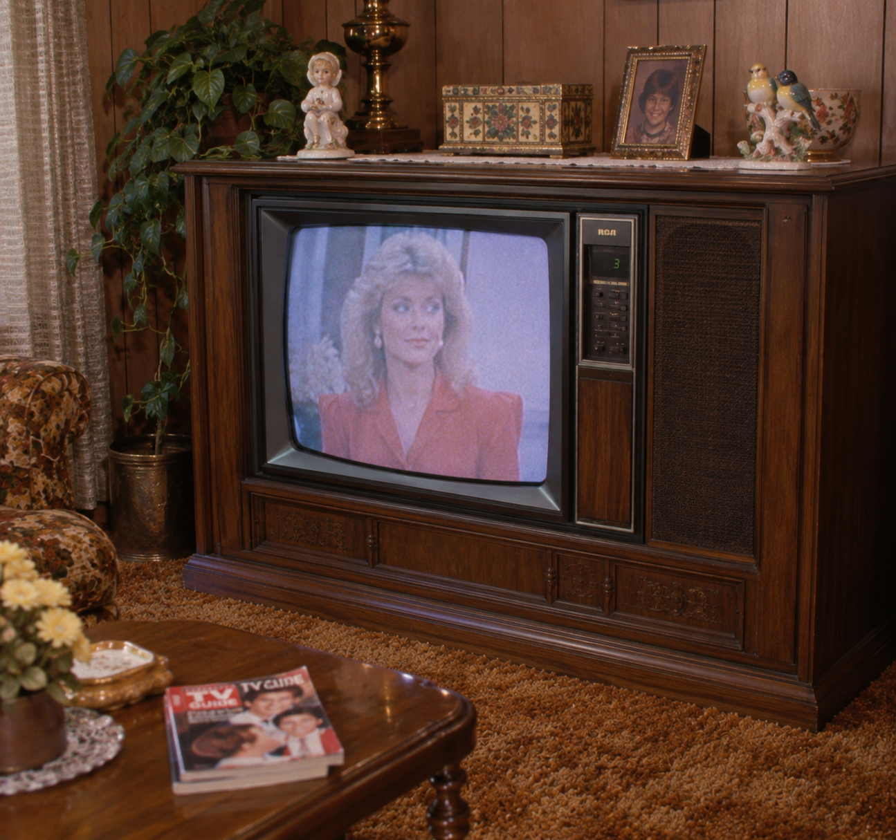 A vintage console TV displays a woman with blonde hair in a red blazer. The TV sits in a wood-paneled living room with framed photos, magazines, plants, and decor, capturing a retro 1970s-80s vibe.