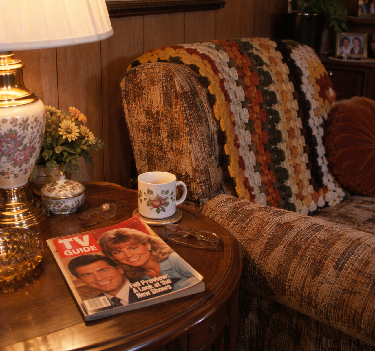A cozy armchair with a multicolored crocheted blanket beside a wooden table holding a lamp, mug, glasses, and a TV Guide magazine featuring two smiling people on the cover.
