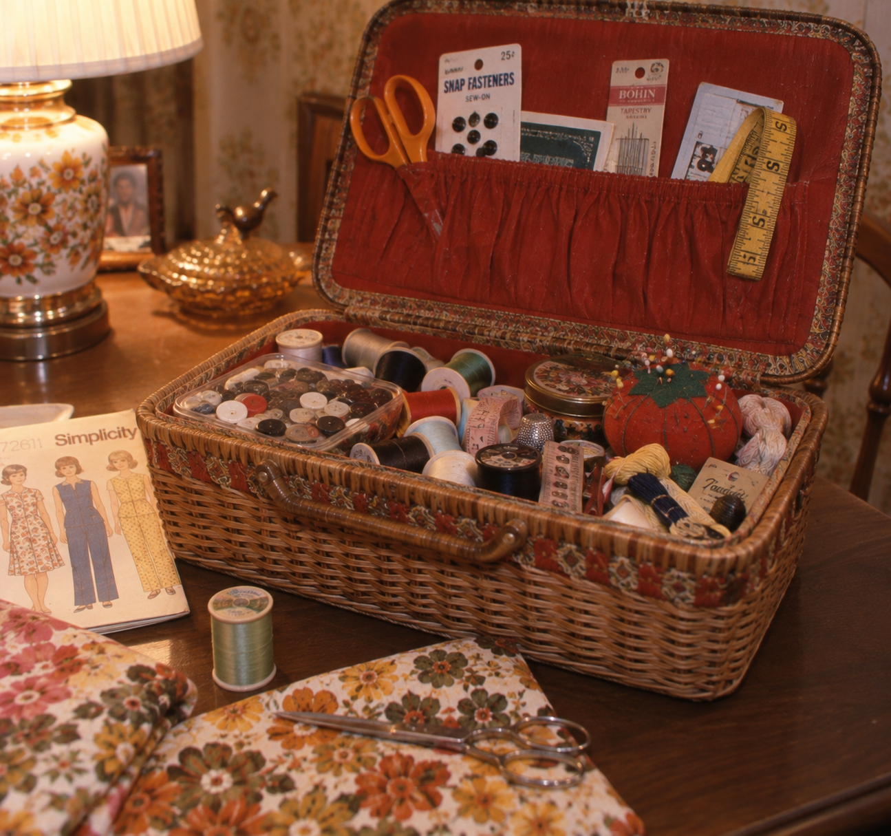 A vintage sewing basket holds thread spools, scissors, a pincushion, and sewing notions. Nearby are floral fabrics, a pattern booklet with dress designs, and a lamp with a floral base on a wooden table.