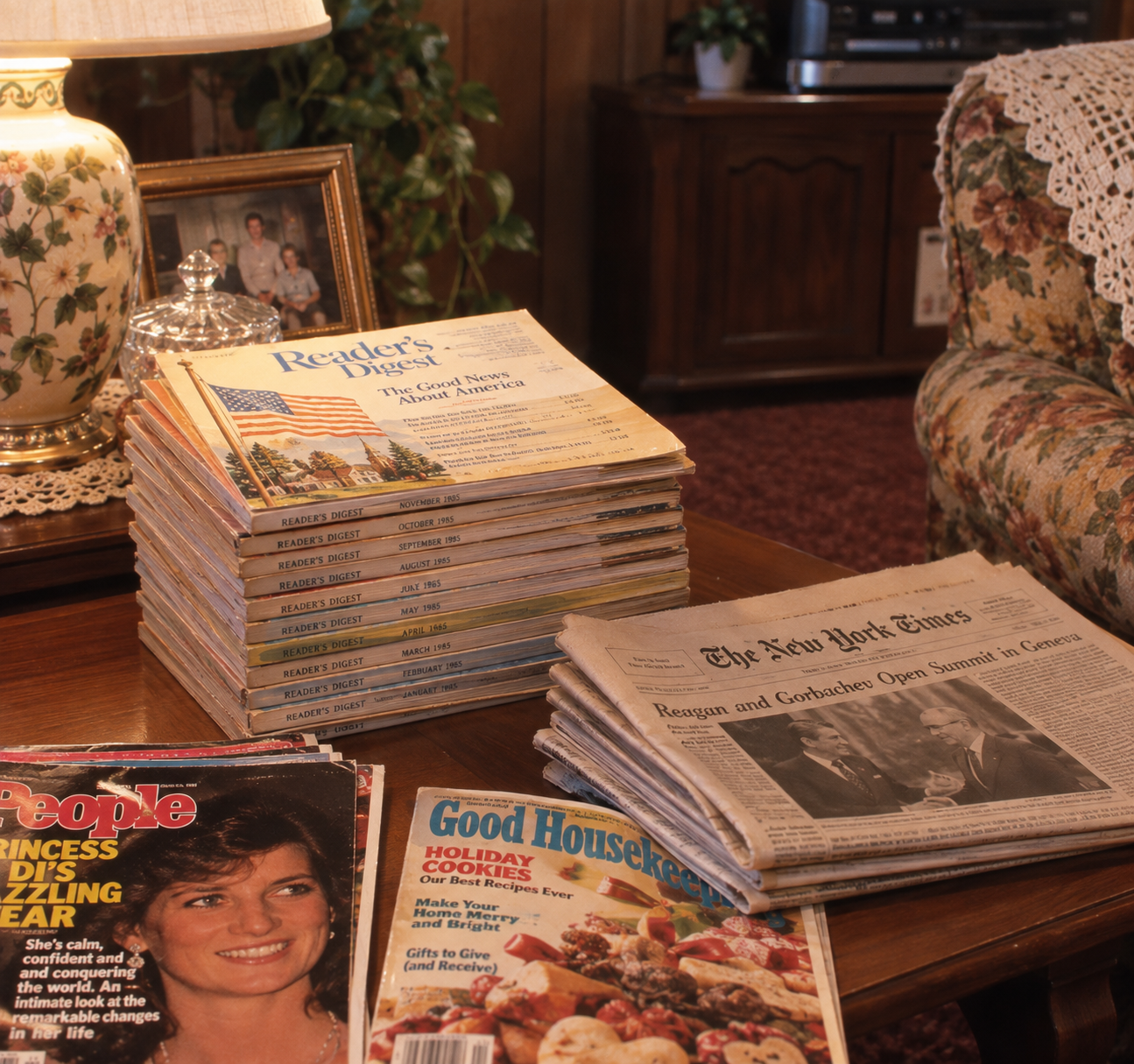 A stack of Reader’s Digest magazines, newspapers, and People and Good Housekeeping magazines sit on a wooden table in a living room beside a lamp, framed photo, and floral couch.