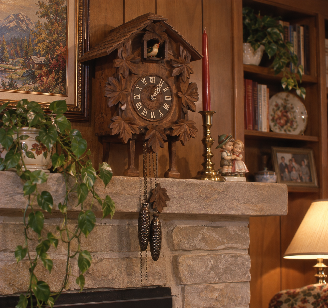 A wooden cuckoo clock hangs above a stone fireplace mantle, decorated with plants, a red candle, figurines, and bookshelves. A lamp and framed art are also visible in this cozy living room scene.