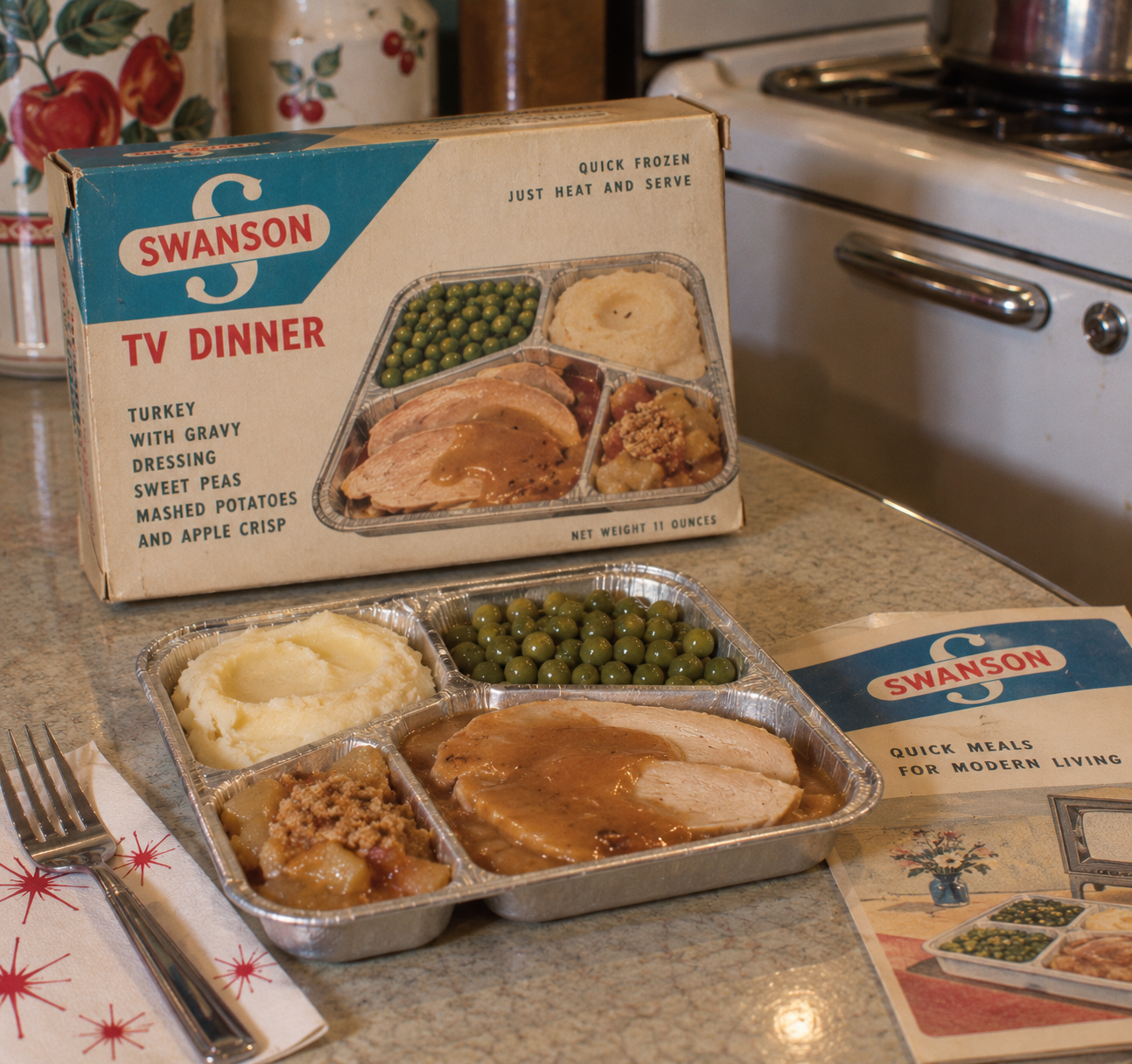 A vintage Swanson TV dinner with turkey, peas, mashed potatoes, stuffing, and dessert sits on a counter, next to its box, a fork, and an illustrated instruction pamphlet. American homes in the 1950s. 