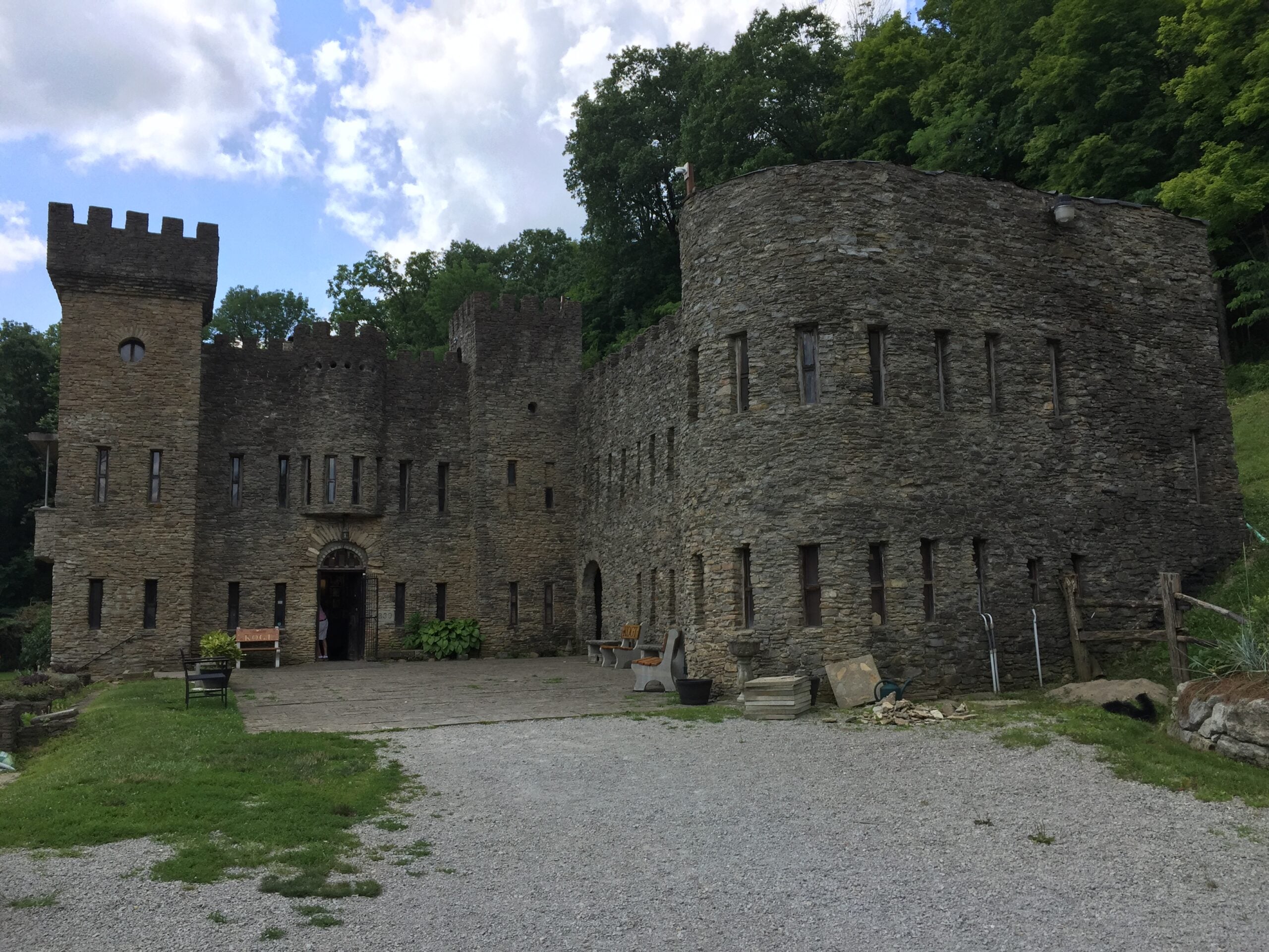 A majestic stone castle with imposing towers and arched windows, surrounded by trees and greenery, sits beside a gravel driveway under a partly cloudy sky, capturing the timeless allure of historic castles.