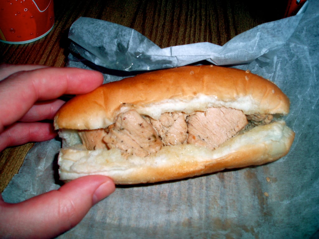A hand holding a hoagie roll filled with chunks of cooked meat, showcasing one of the restaurant specialties, rests on wax paper on a wooden table.