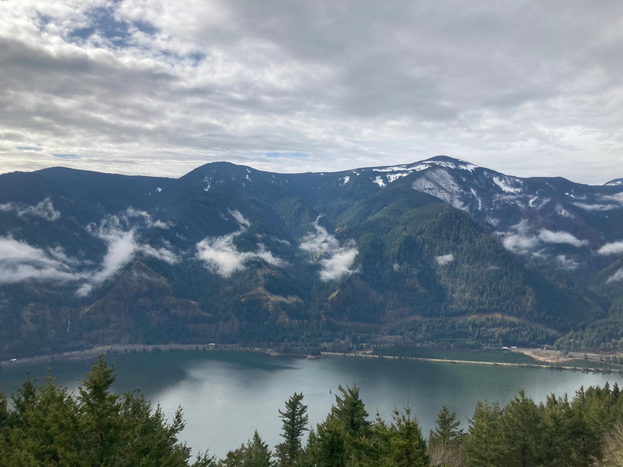 Clouds drift over forested mountains with patches of snow, reflected in a calm lake below. Pine trees frame the foreground under a partly cloudy sky.