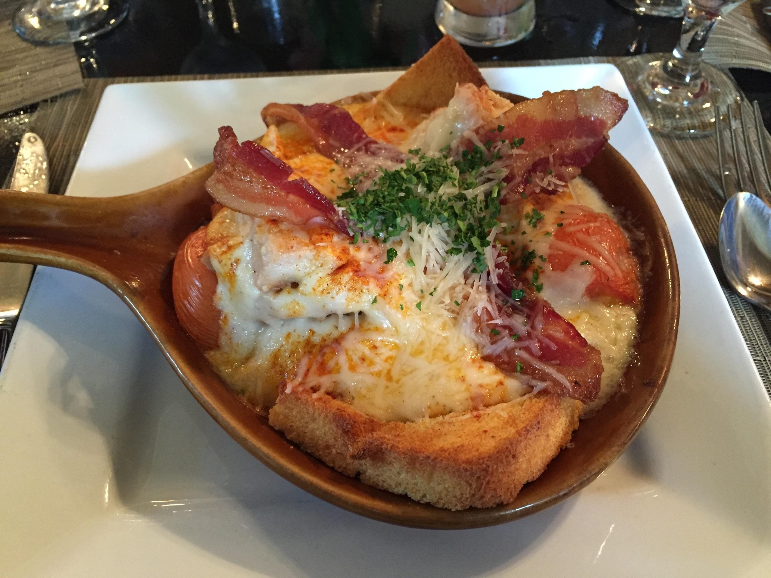 A baked dish in a brown ceramic bowl, showcasing one of the restaurant specialties, features toast, melted cheese, bacon strips, tomato slices, and chopped herbs, served on a white plate with a spoon beside it.