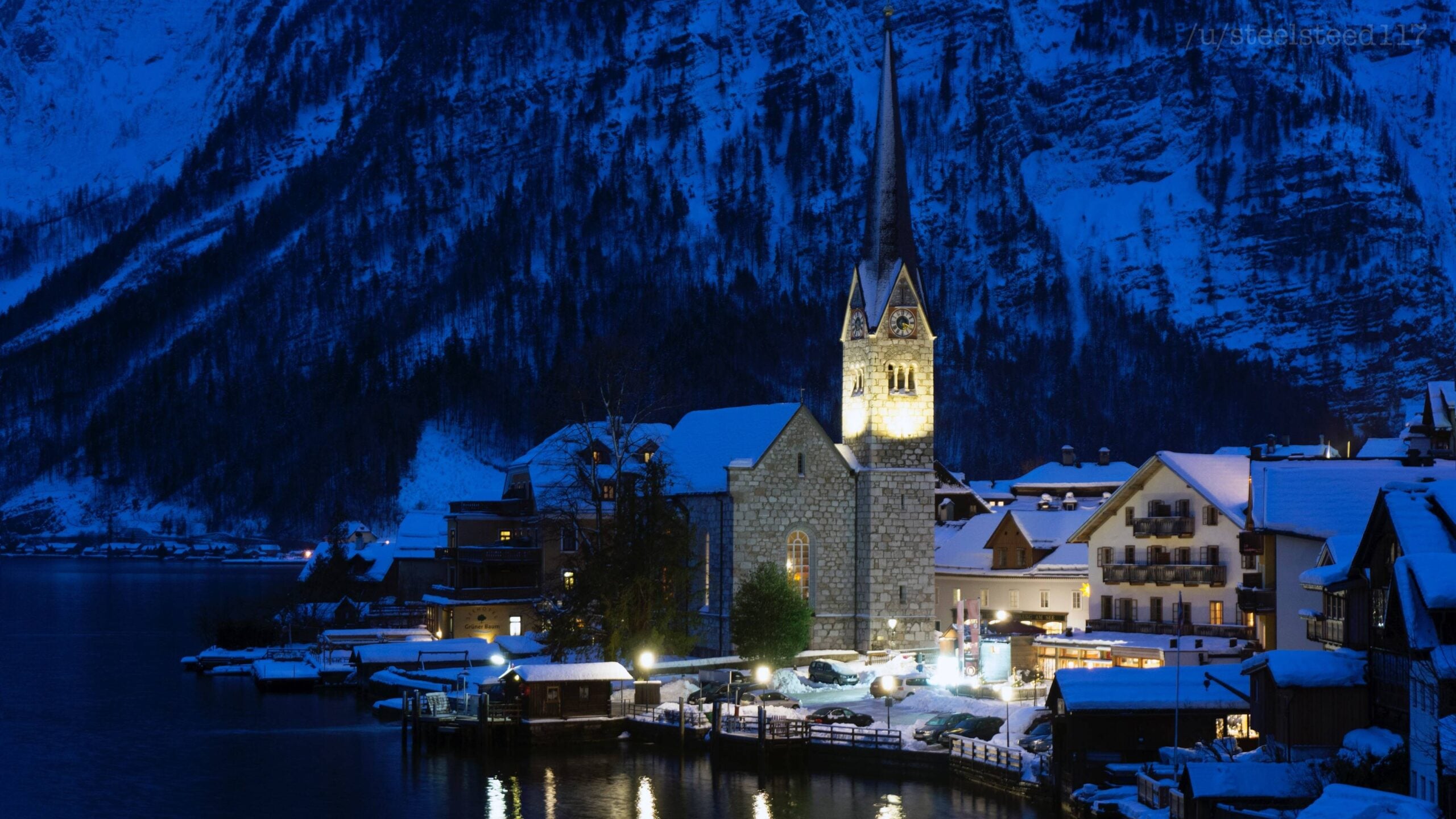 Snow-covered village at dusk with a stone church, tall steeple, and warm lights glowing from buildings, nestled beside a calm lake and surrounded by dark, snowy mountains.