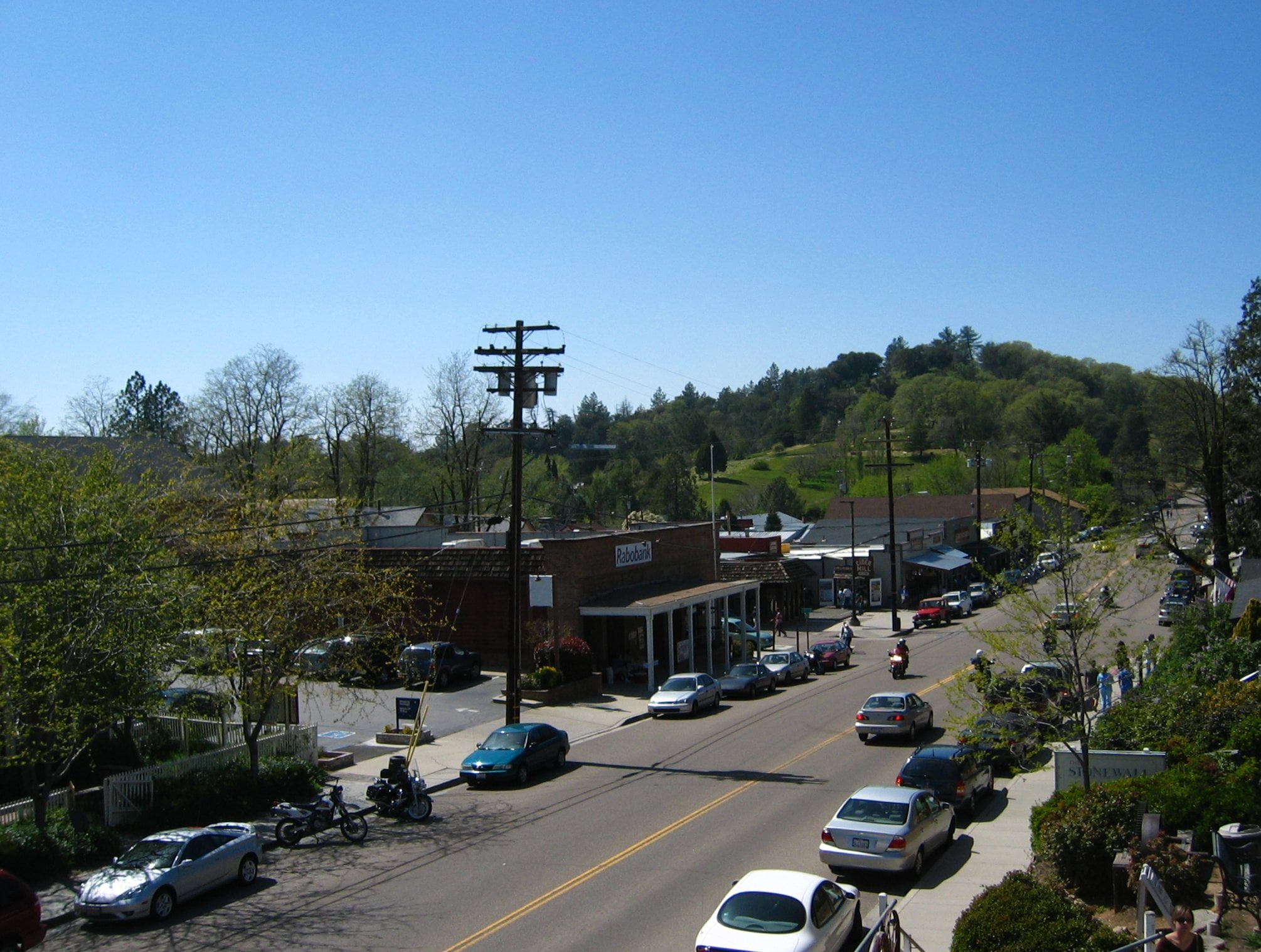 A small town street with parked cars, a few pedestrians, and brick buildings. Trees and hills are visible in the background under a clear blue sky.