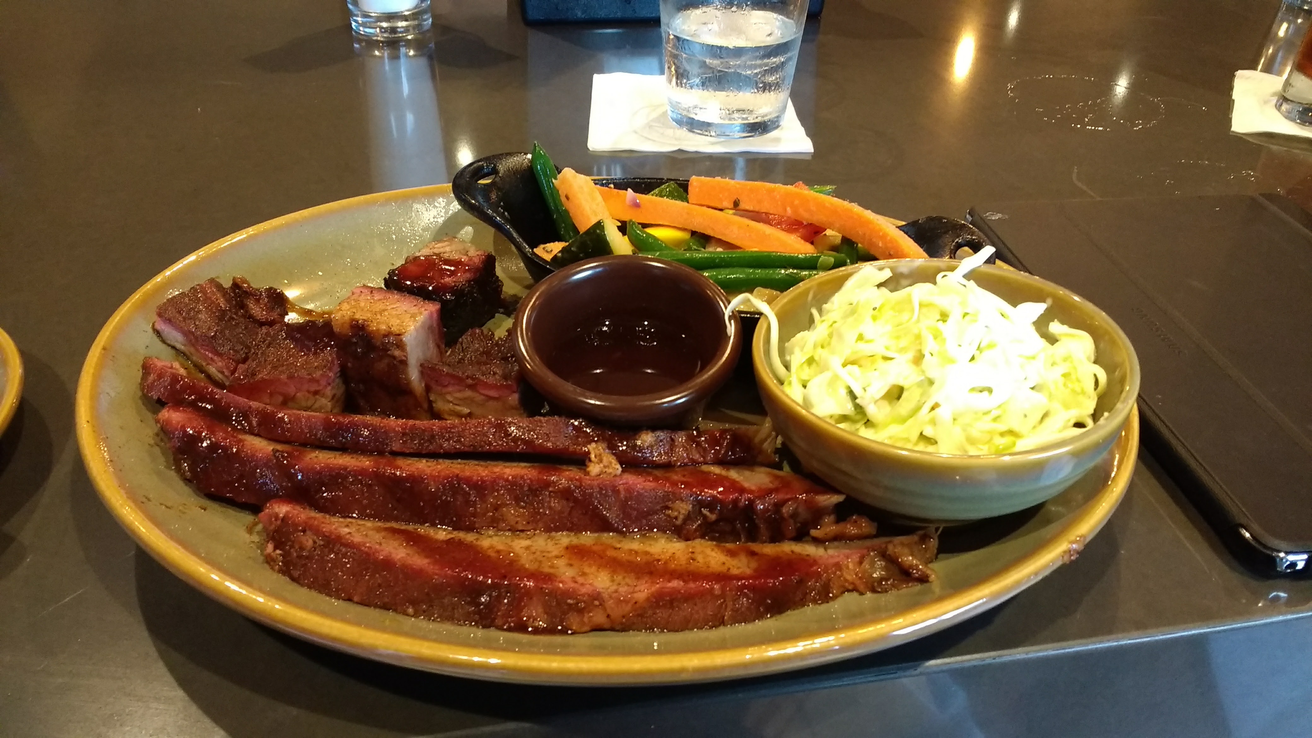 A plate of BBQ featuring sliced brisket, coleslaw, cooked carrots and green beans, and a ramekin of BBQ sauce—classic flavors you’ll find in the best American food towns.
