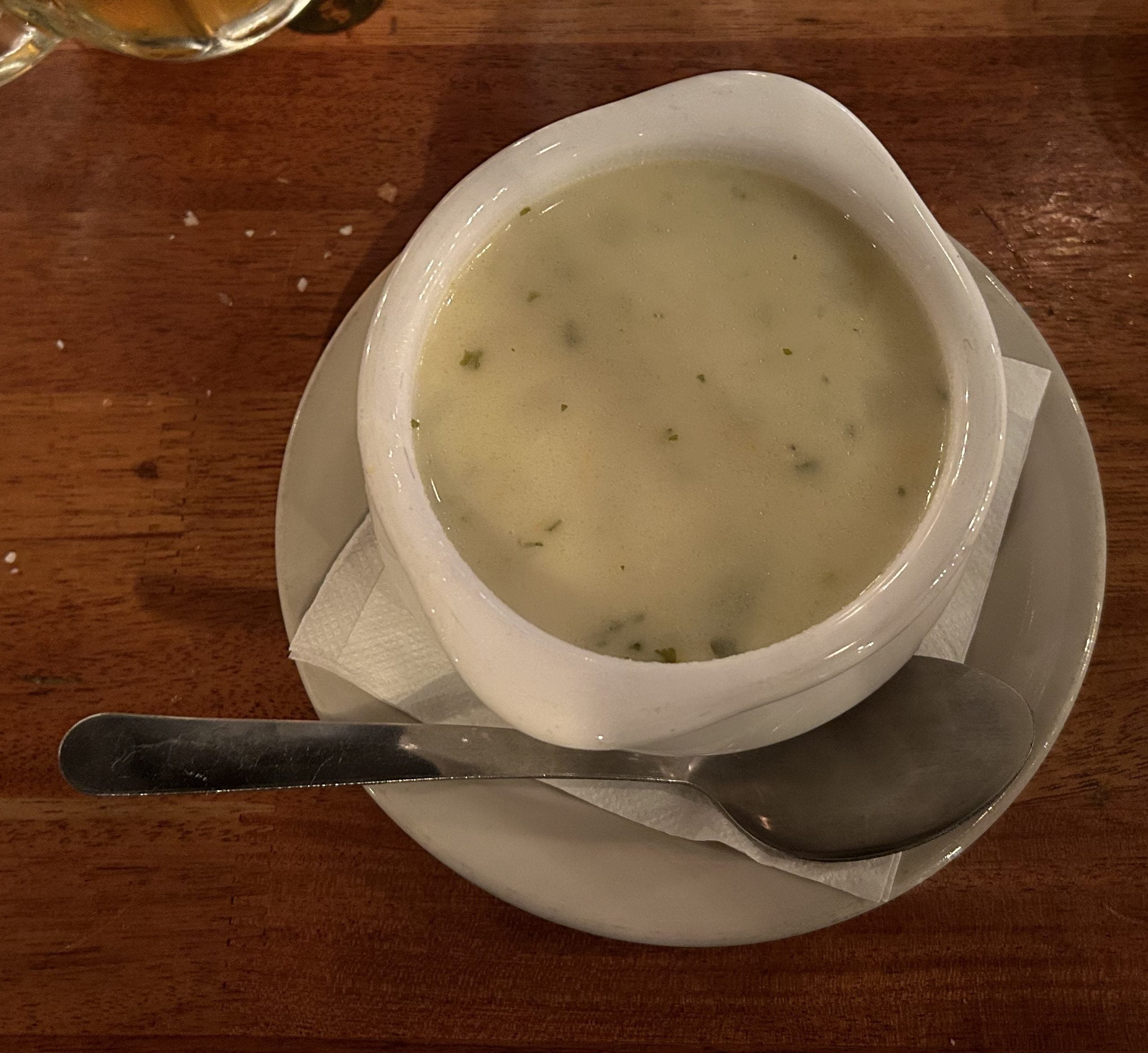 A white bowl of creamy soup, one of the restaurant specialties, is garnished with herbs and sits on a saucer with a spoon beside it, placed on a wooden table.