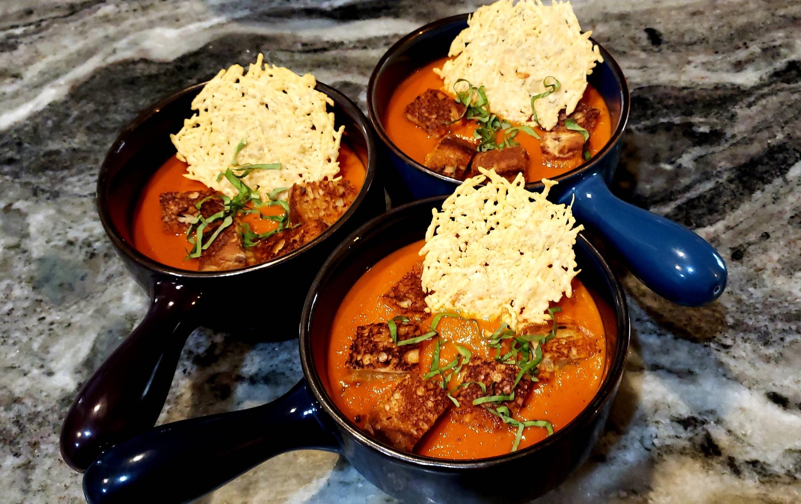 Three bowls of tomato soup topped with croutons, shredded basil, and a large parmesan crisp, arranged on a marbled countertop—perfectly paired with classic grilled cheese sandwiches for the ultimate comforting meal.