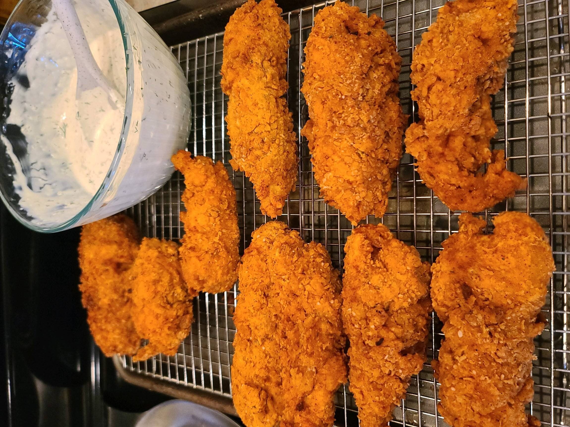 Golden fried chicken tenders rest on a wire rack beside a bowl of creamy white dipping sauce, likely ranch, with herbs, on a dark baking sheet.