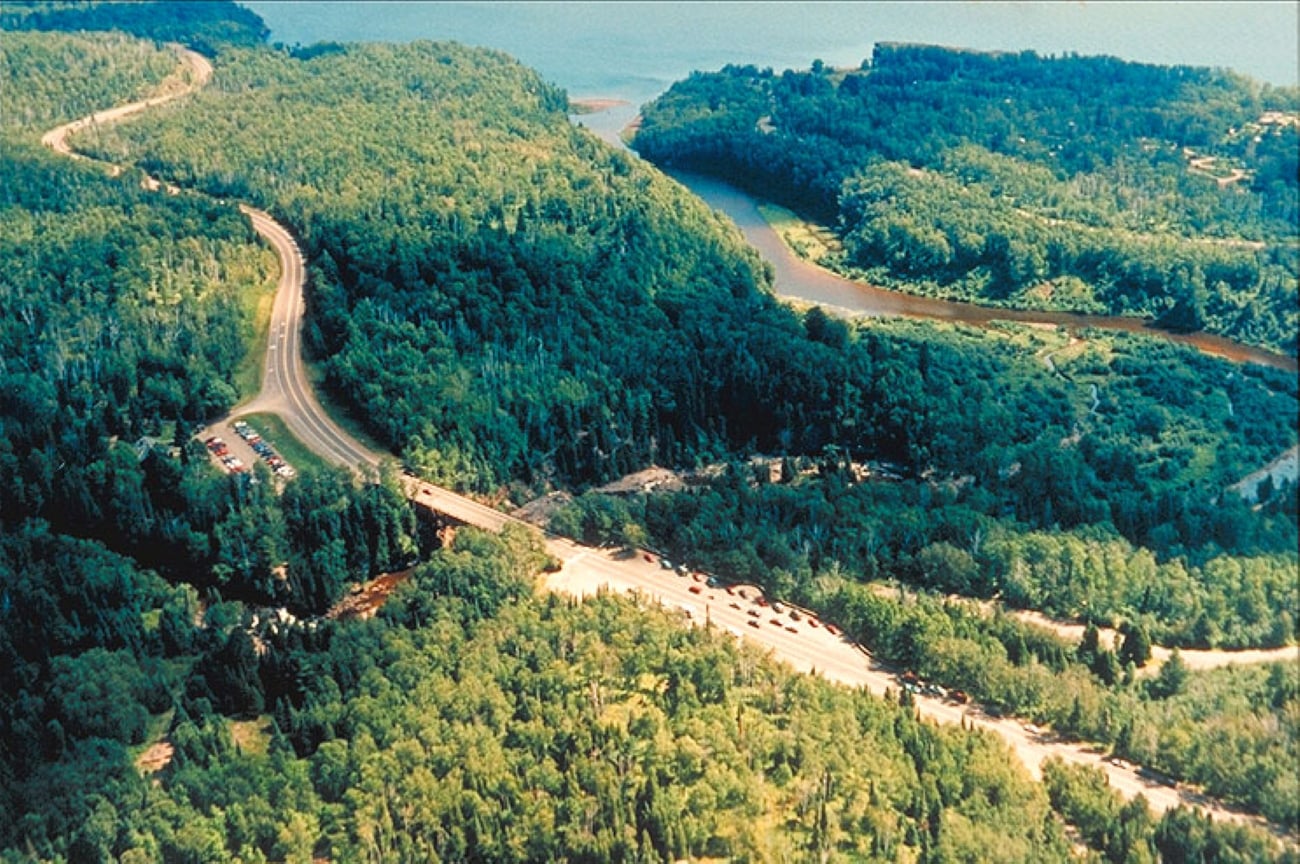 Aerial view of a forested landscape with winding roads—perfect for trips where getting there is part of the adventure—a river crossing under a bridge and vehicles amid dense green trees.