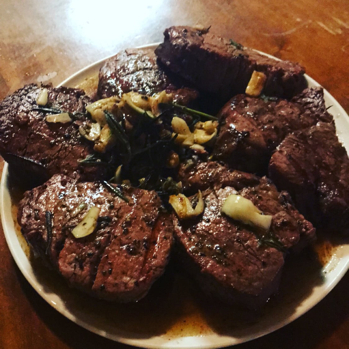 A plate of cooked steak medallions garnished with sliced garlic and herbs, served on a wooden table.