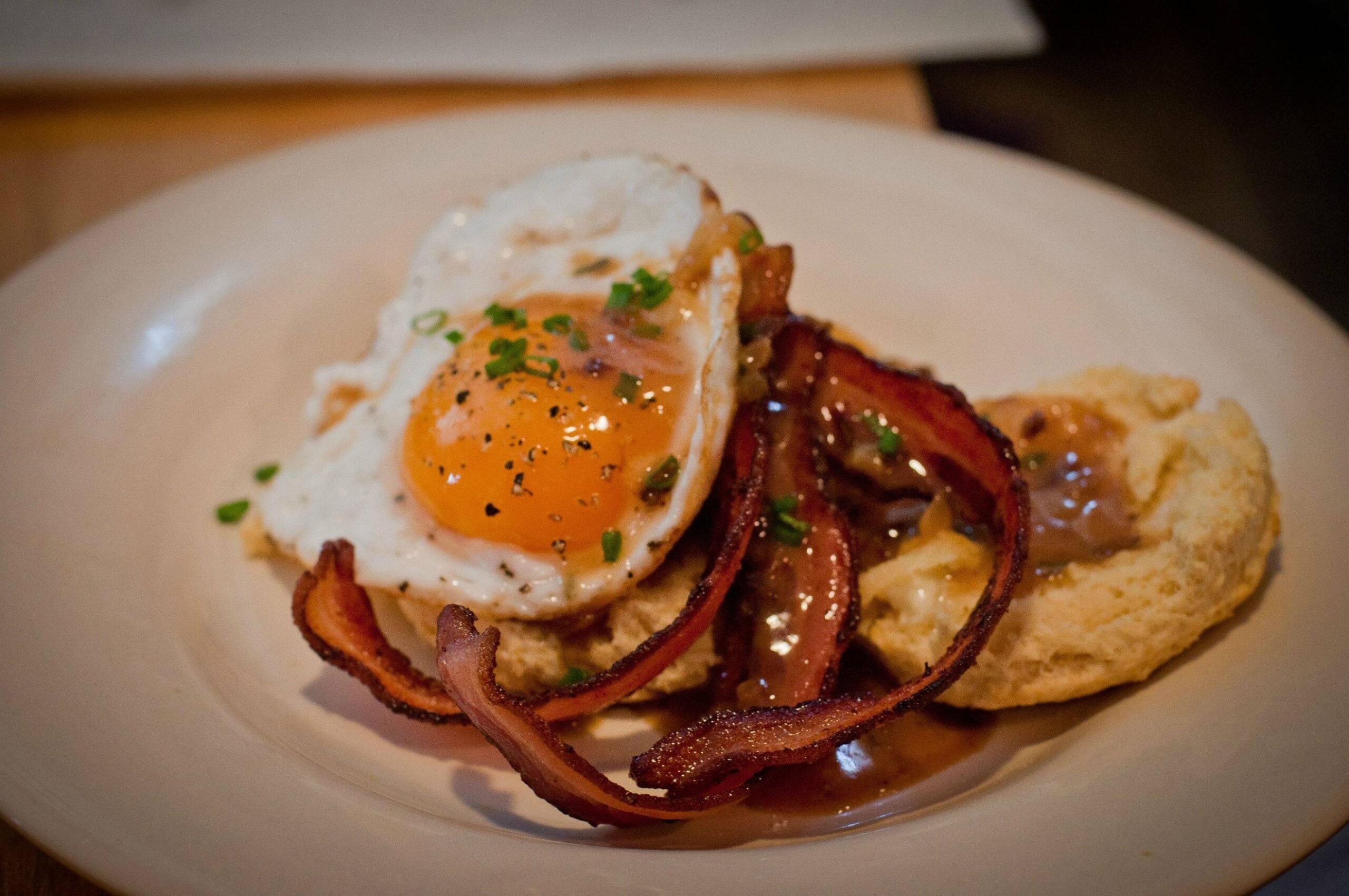 A plate with a biscuit topped with brown gravy, crispy bacon strips, and a sunny-side-up egg garnished with chopped chives.