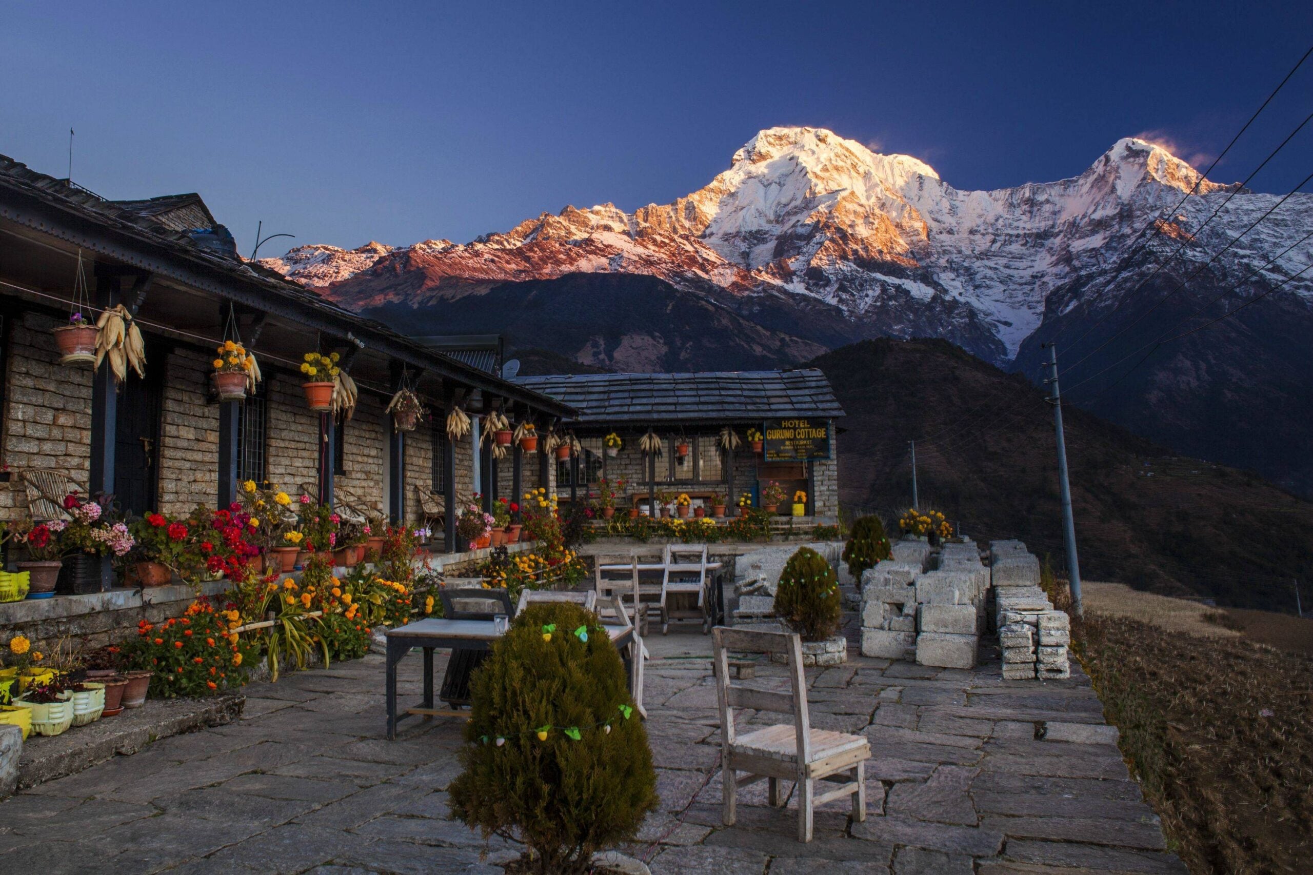 Stone patio with tables and chairs, flower pots, and mountain lodge at sunrise. Snow-capped peaks and blue sky are in the background.