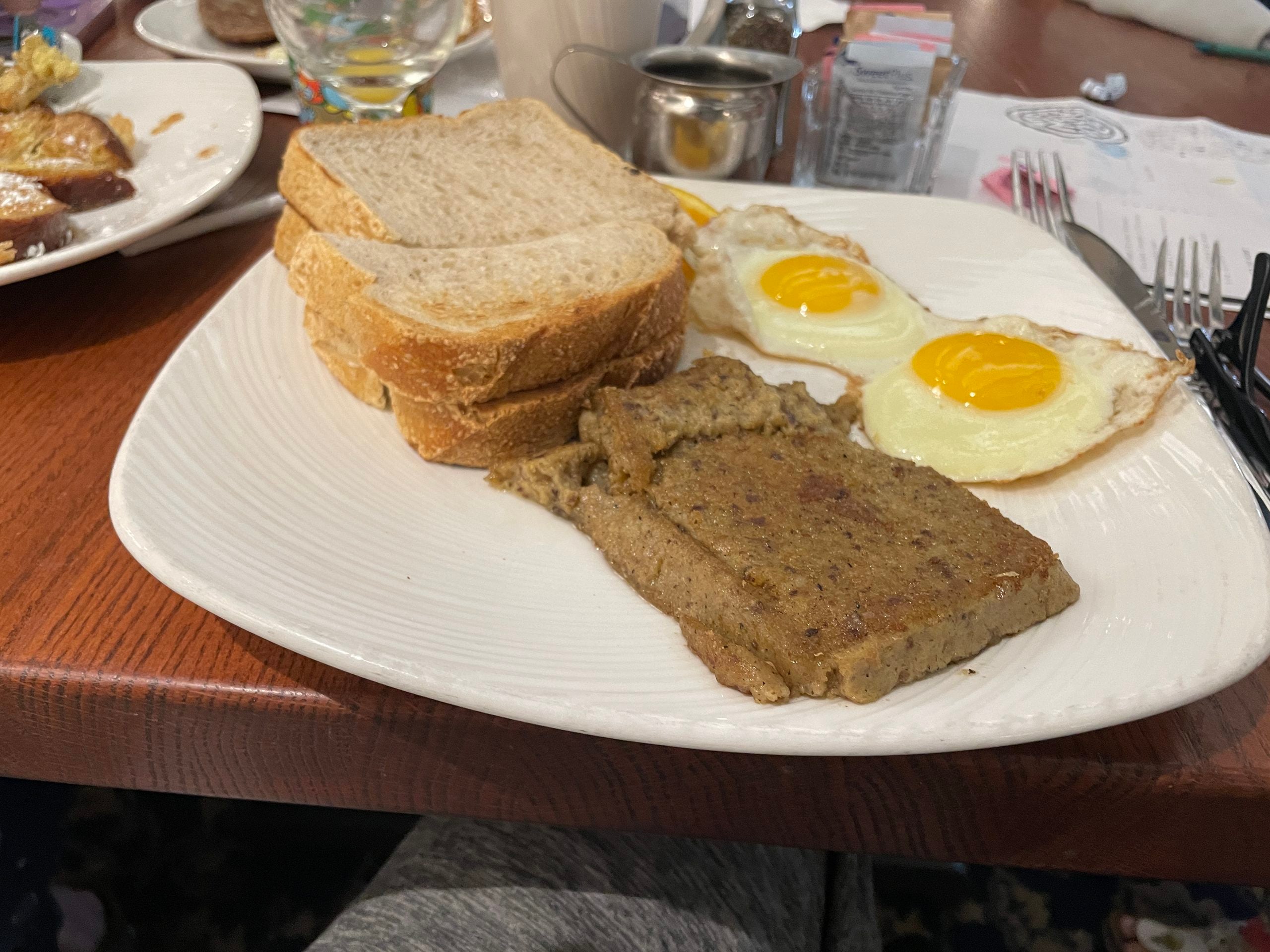 A plate with two sunny-side-up eggs, several slices of white bread, and a slice of scrapple—one of the restaurant specialties—rests on a wooden table at a restaurant. Other food and drinks are partially visible in the background.