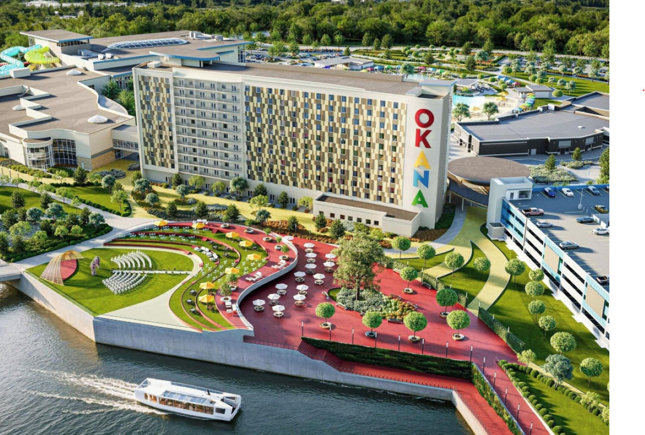 Aerial view of a riverside complex with a modern hotel labeled “OKANA,” outdoor seating, amphitheater, walking paths, greenery, a large parking lot, and a boat on the water in the foreground—offering the inviting atmosphere typically found at premier resorts.
