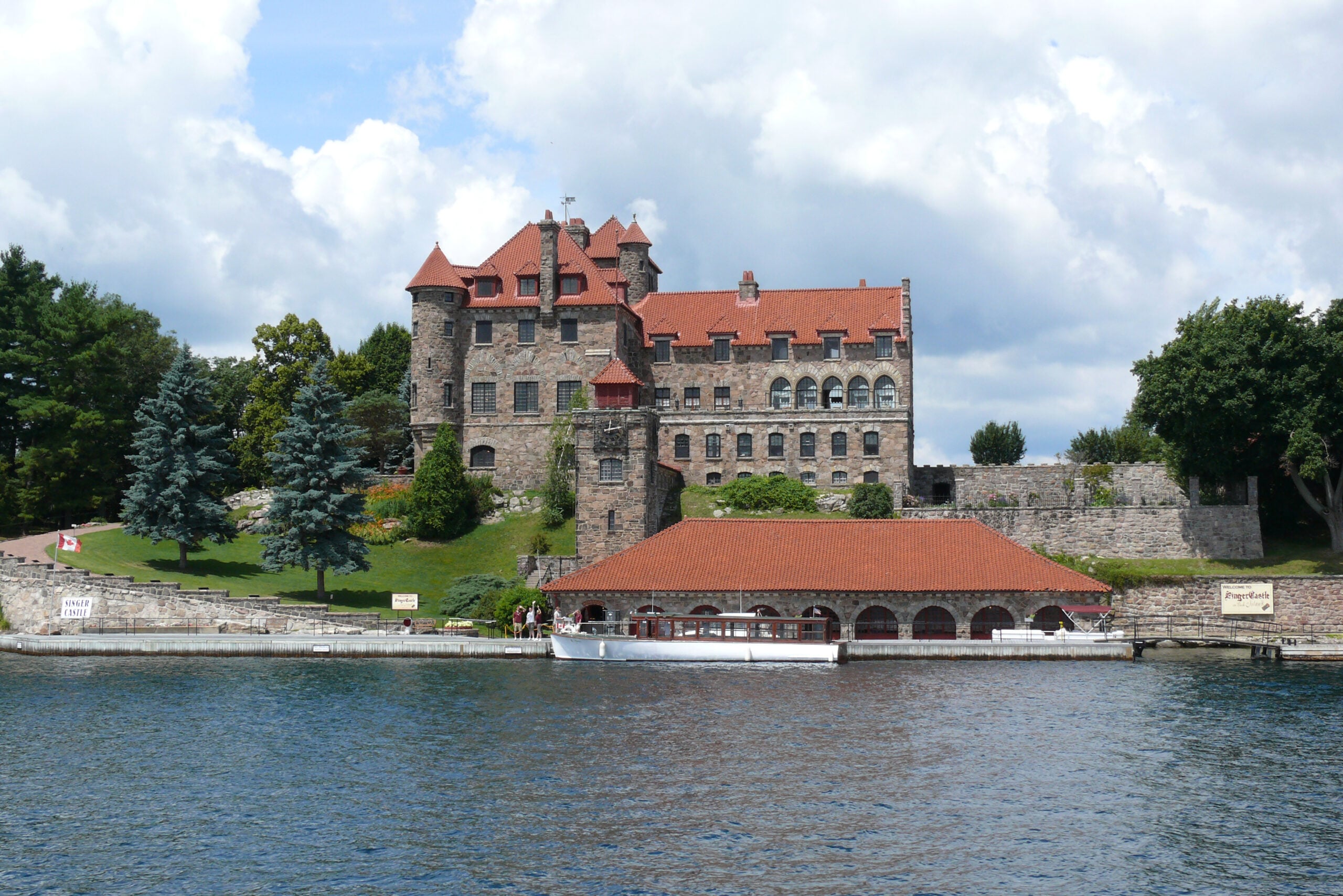 A large stone castle with red roofs sits by the water, surrounded by green trees and a boathouse in the foreground. Like many historic castles, its imposing presence is complemented by the serene setting, while the sky above remains partly cloudy.