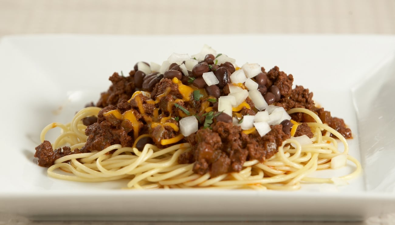 A plate of spaghetti topped with chili, shredded cheddar cheese, black beans, and diced onions—a hearty combination of comfort foods—served on a white square plate.