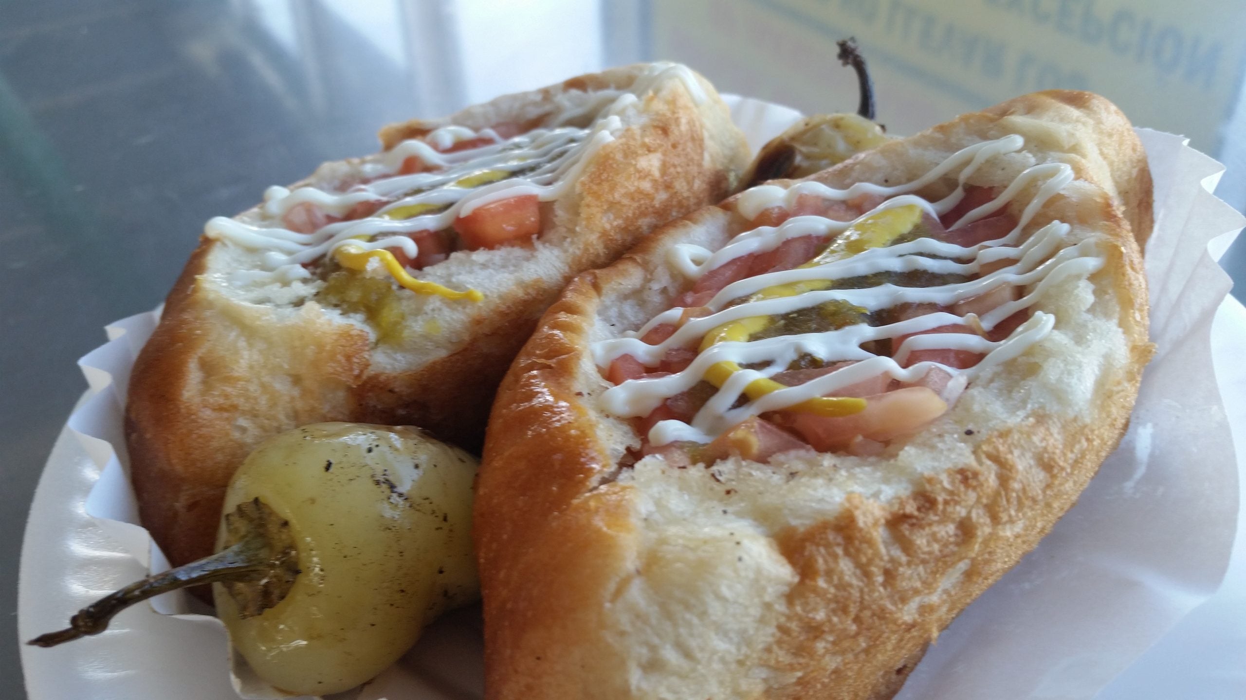 Two hot dog buns filled with sausage, topped with mustard, mayonnaise, diced tomatoes, onions, and jalapeños—one of our restaurant specialties—served on a paper tray with a pickled pepper on the side.