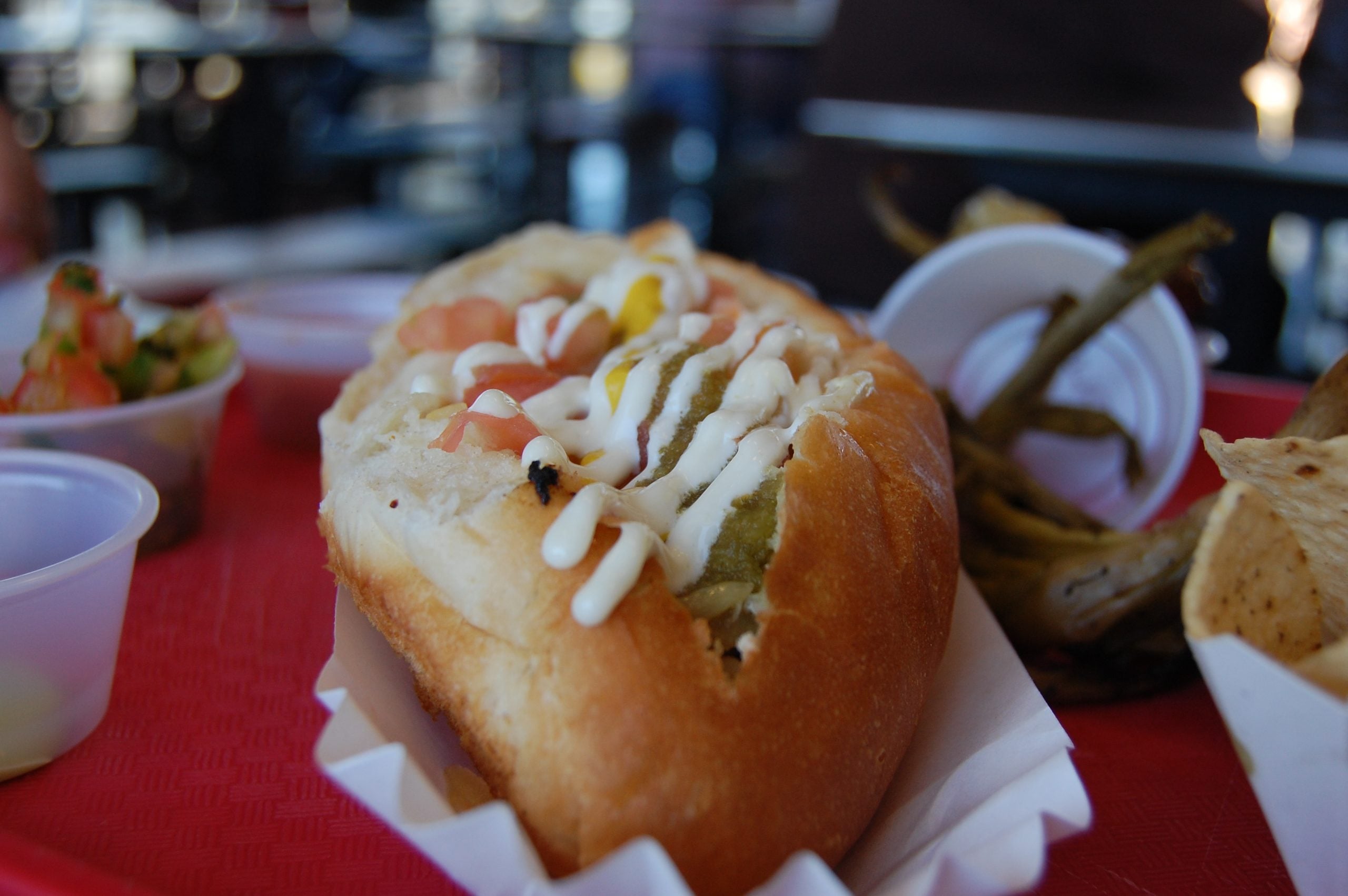 A close-up of a sandwich topped with diced vegetables and mayonnaise on a red tray, evoking the flavors found in American food towns, with pickled green beans, chips, and salsa in the background.