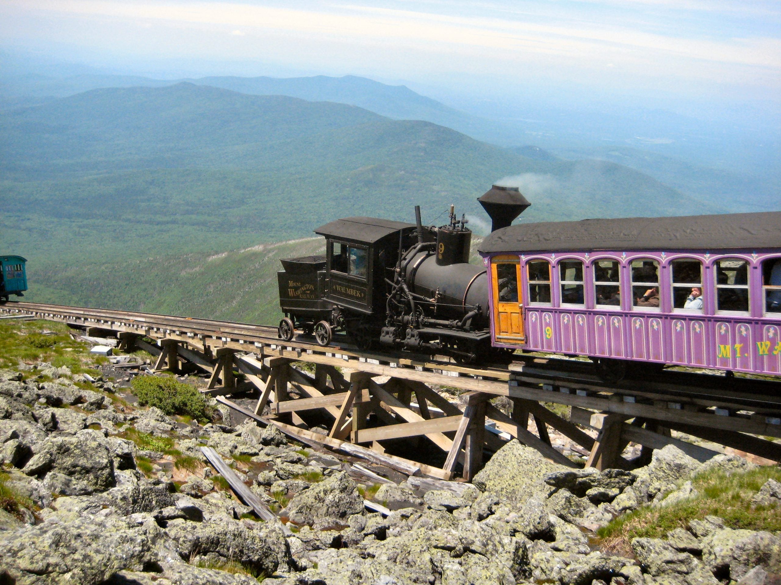 A black steam train pulls colorful passenger cars up a steep, rocky mountain track—proof that some trips where getting there is half the adventure—set against green hills and a hazy blue sky.